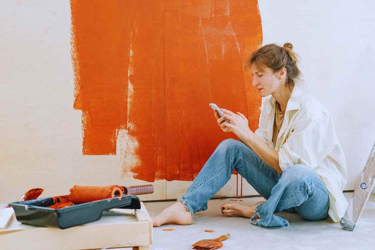 Donna seduta a terra accanto ad una parete dipinta solo in parte che guarda il telefono e attrezzi per la pittura poggiati su un tavolino