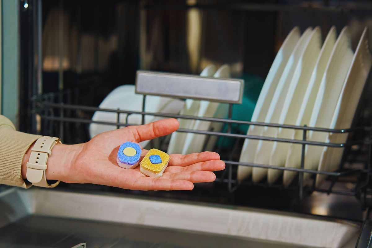 Persona che tiene sul palmo della mano due pastiglie per lavastoviglie davanti ad una lavastoviglie carica.