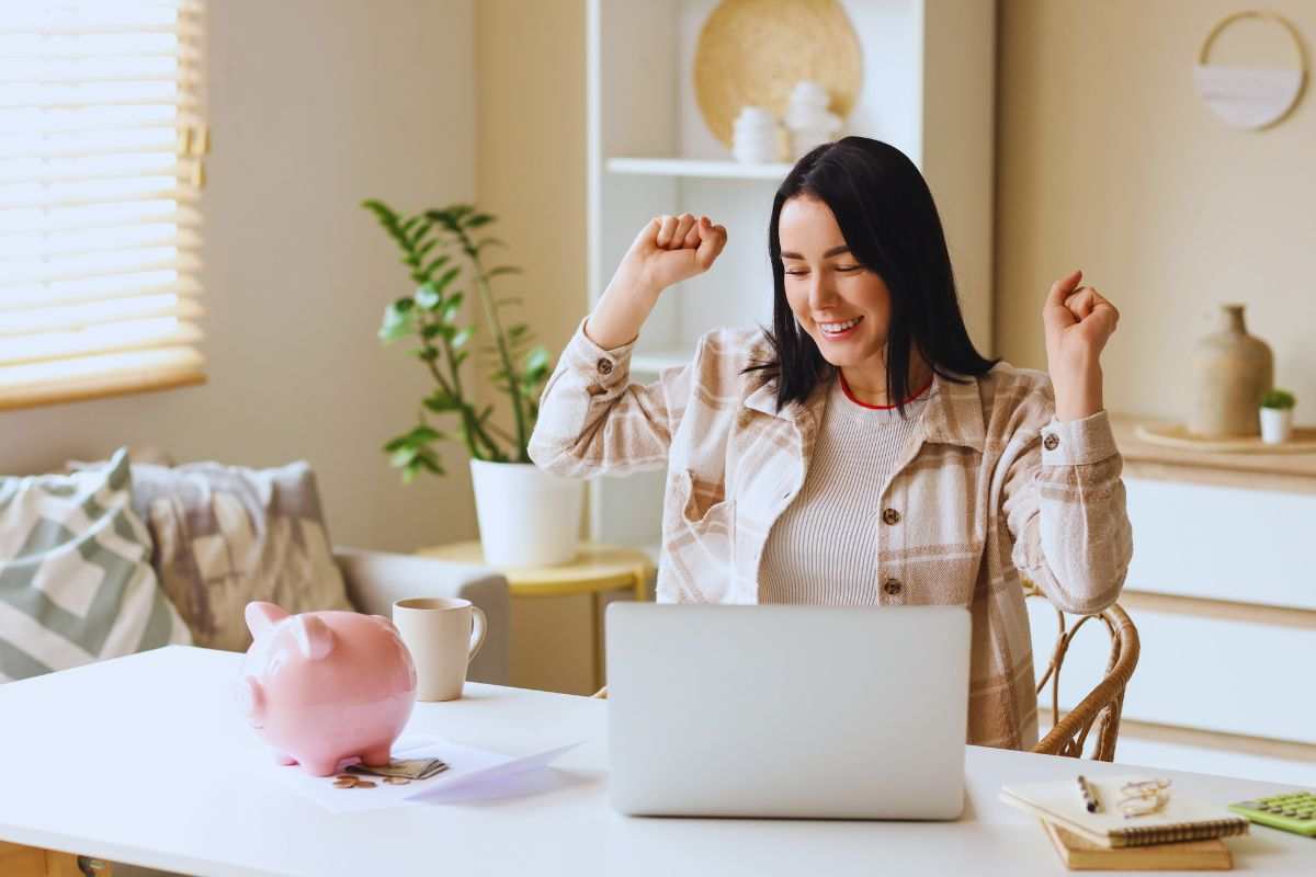 Ragazza seduta alla scrivania che esulta - salvadanaio e portatile poggiati su tavolo