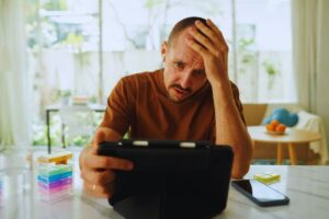 Uomo seduto al tavolo della cucina di casa che tiene in mano un tablet e con l'altra si tiene la fronte