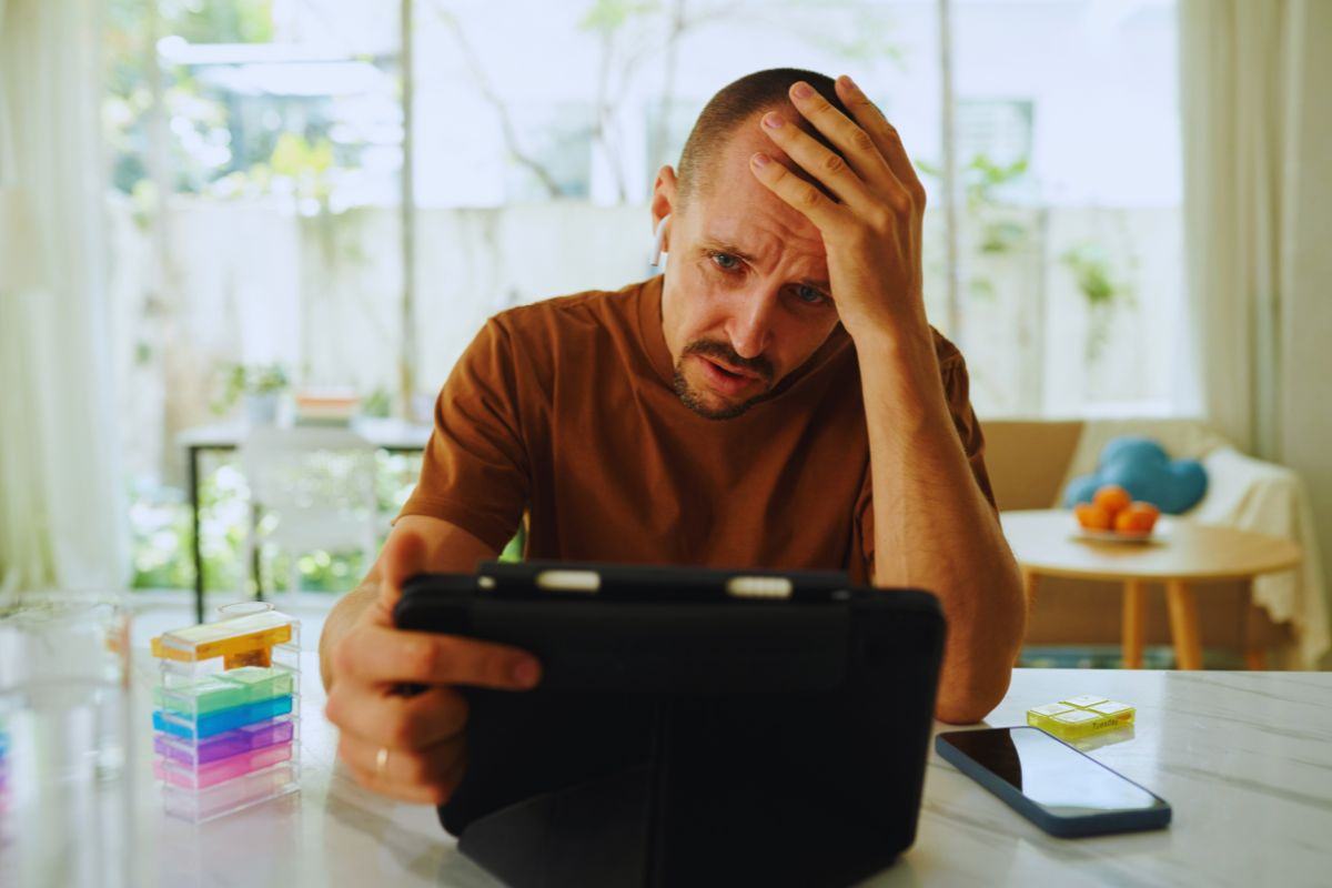 Uomo seduto al tavolo della cucina di casa che tiene in mano un tablet e con l'altra si tiene la fronte