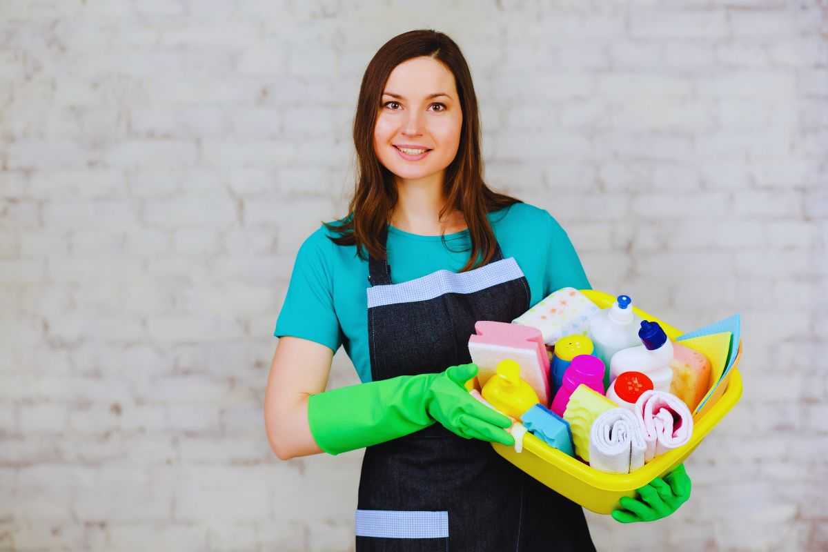 Donna con indosso grembiule e guanti che tiene in mano una cesta di prodotti per la pulizia della casa