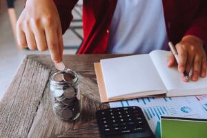 Ragazzo che mette una moneta in un barattolo utilizzato come salvadanaio e nel frattempo fa i conti su un taccuino